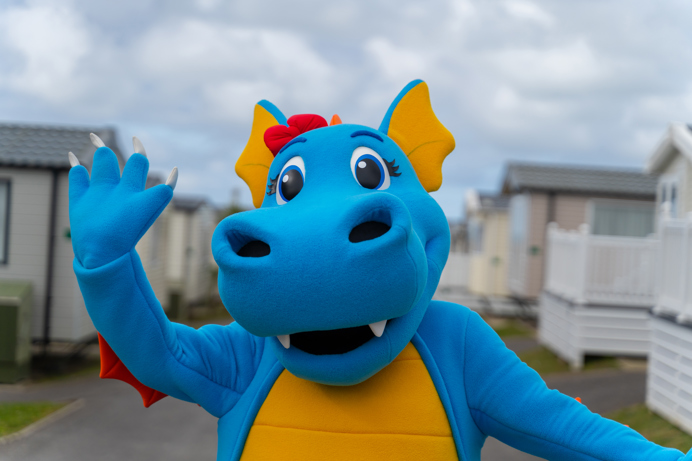 A dinosaur mascot, Vic, walking through a Chesil Beach Holiday Park waving at the camera 