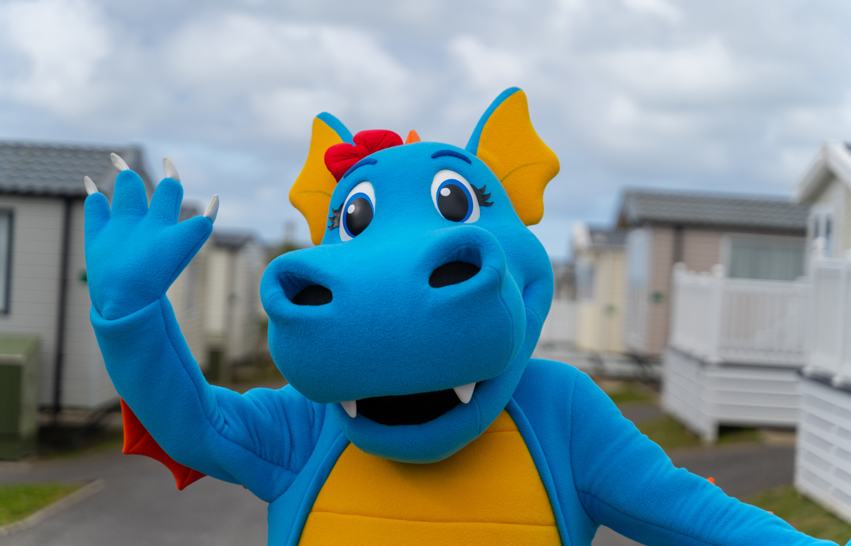 A dinosaur mascot, Vic, walking through a Chesil Beach Holiday Park waving at the camera 