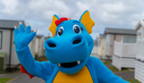 A dinosaur mascot, Vic, walking through a Chesil Beach Holiday Park waving at the camera 