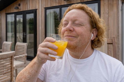 A man happily drinking orange juice on the decking of a luxury lodge while listening to music with his headphones in