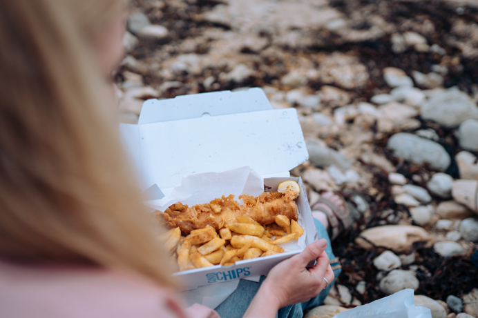 A woman sat on the beach with a fish and chip box open on her lap 