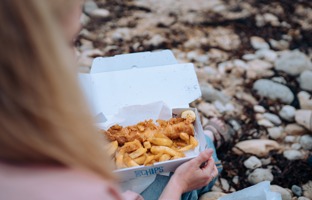 A woman sat on the beach with a fish and chip box open on her lap 