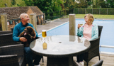 A couple sat at a table outside on a terrace overlooking an outdoor pool with a dog