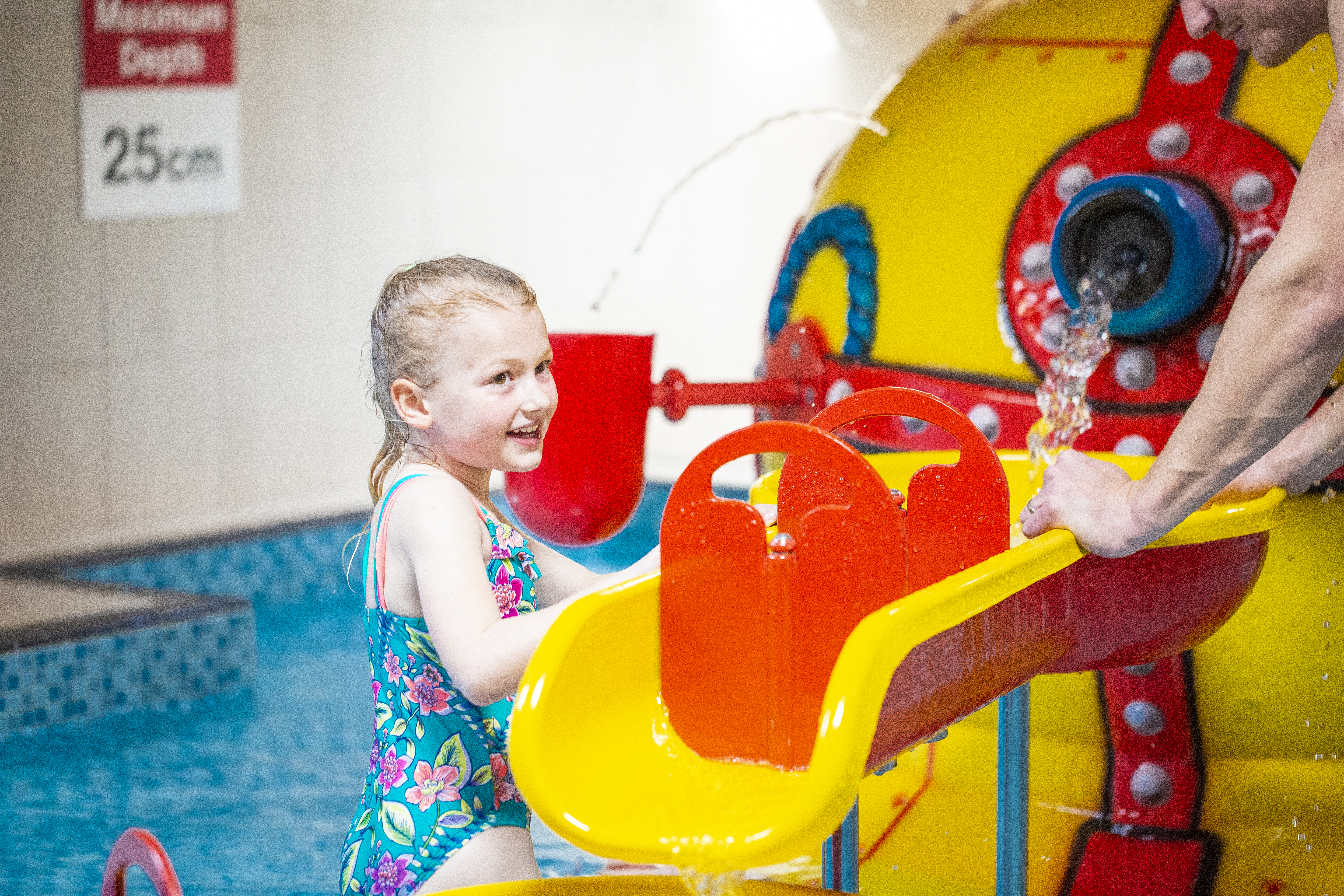 A smiling young girl with an adult beside her playing in a shallow splash pool