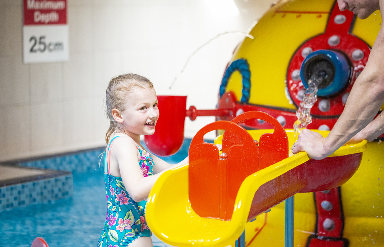 A smiling young girl with an adult beside her playing in a shallow splash pool