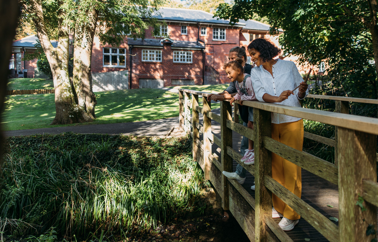 A family of three on a wooden bridge looking over a stream beside a country club restaurant building surrounded by woodland