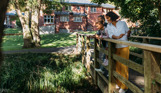 A family of three on a wooden bridge looking over a stream beside a country club restaurant building surrounded by woodland