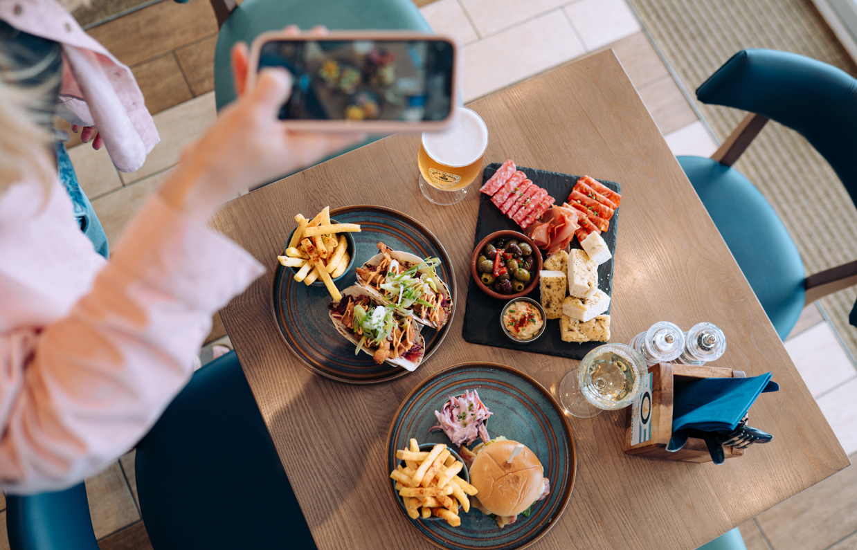 A selection of dishes including steak, burger, tacos, charcuterie board, taken from above with a woman taking a photo also from above with her iPhone