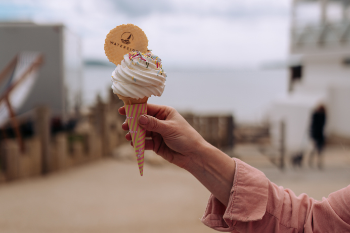 A Mr Whippy style ice cream in a cone with a Waterside wafer being held up against the backdrop of the sea