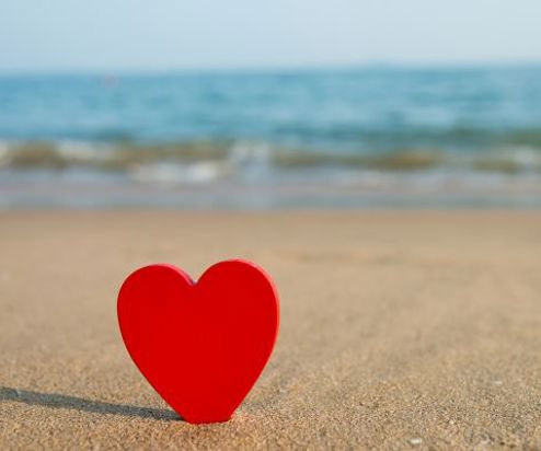 A red heart stood upright in the sand with the sea in the background