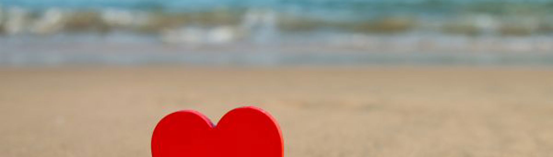 A red heart stood upright in the sand with the sea in the background