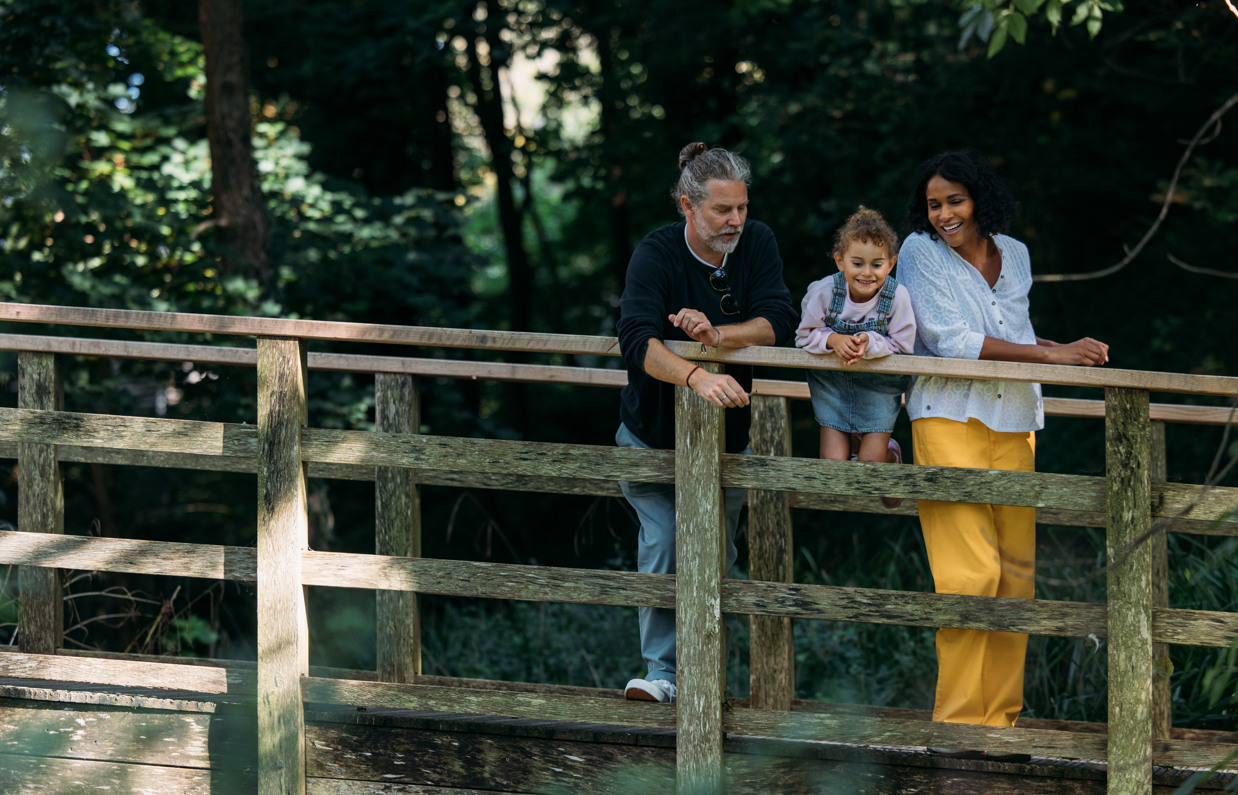 A family of three on a wooden bridge overlooking a stream surrounded by woodland