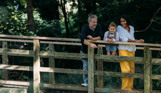 A family of three on a wooden bridge overlooking a stream surrounded by woodland
