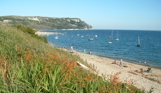 A shingle beach with calm sea, rocky cliffs and various boats on a sunny blue sky day