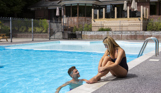 A country club with a large heated outdoor pool among countryside on a blue sunny day with a couple sat in and beside the pool looking at each other