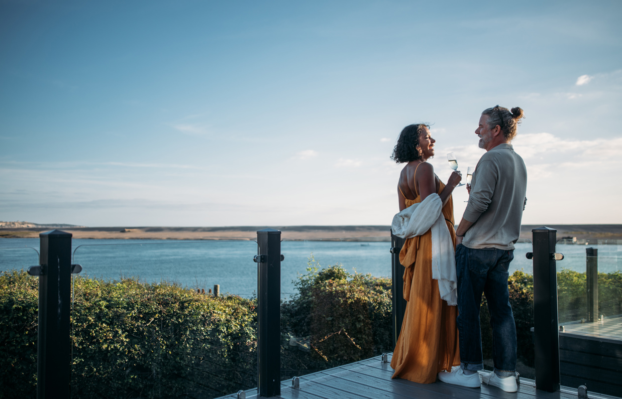 Couple toasting with sparkling wine at dusk on the deck of their lodge overlooking Chesil beach
