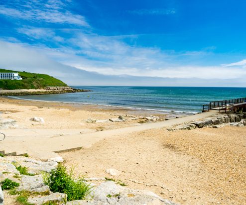 A quiet shingle beach with grassy headland, a small foot pier and calm waters