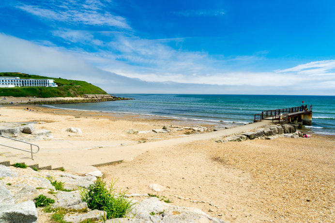 A quiet shingle beach with grassy headland, a small foot pier and calm waters