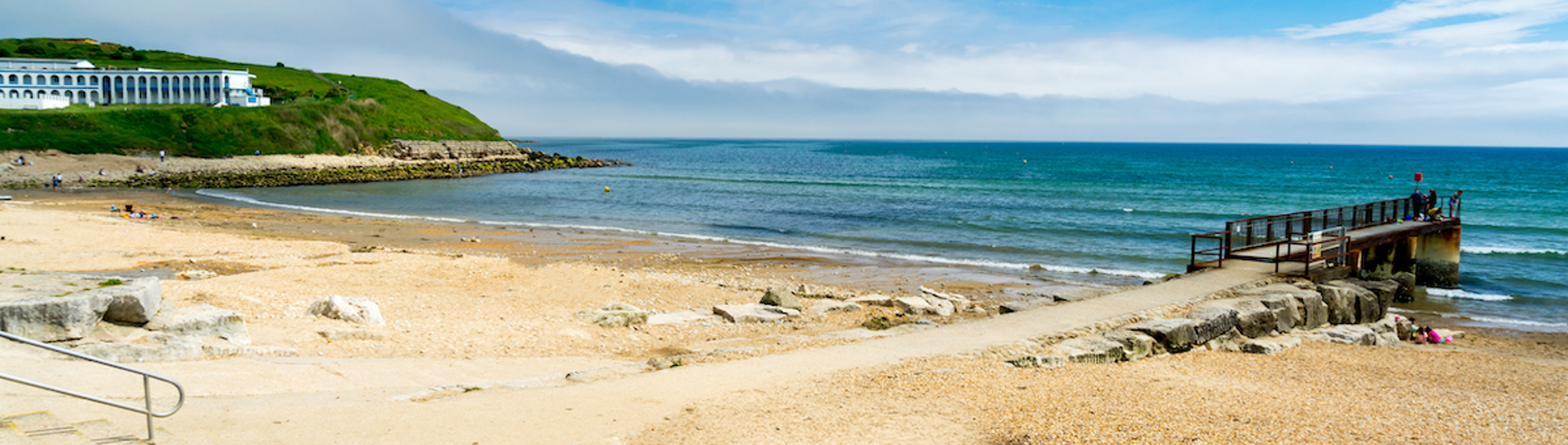 A quiet shingle beach with grassy headland, a small foot pier and calm waters