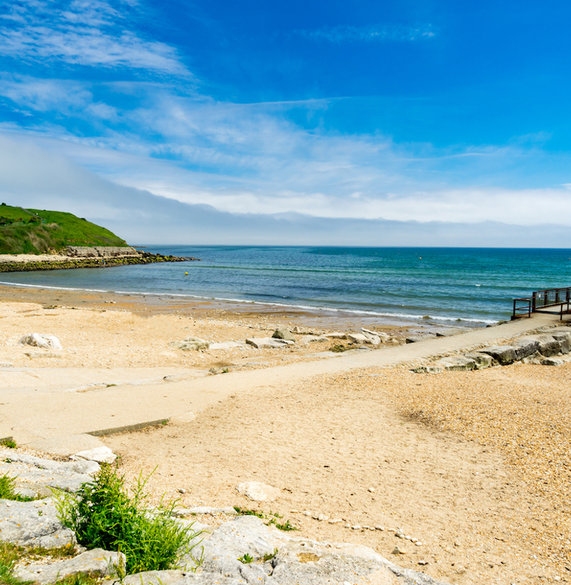 A quiet shingle beach with grassy headland, a small foot pier and calm waters