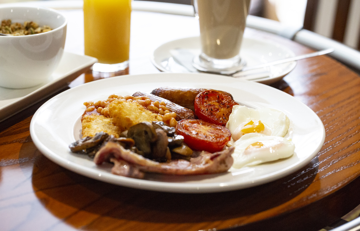A full English breakfast on a table with other breakfast items such as cereal, orange juice and coffee