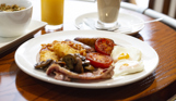 A full English breakfast on a table with other breakfast items such as cereal, orange juice and coffee