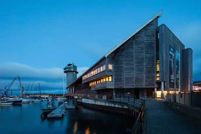 The outside of a striking building by the water at dusk with the lights on inside