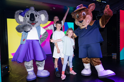 A kangaroo mascot, Boomer, and a koala mascot, Kimi on stage with two young children and an entertainer