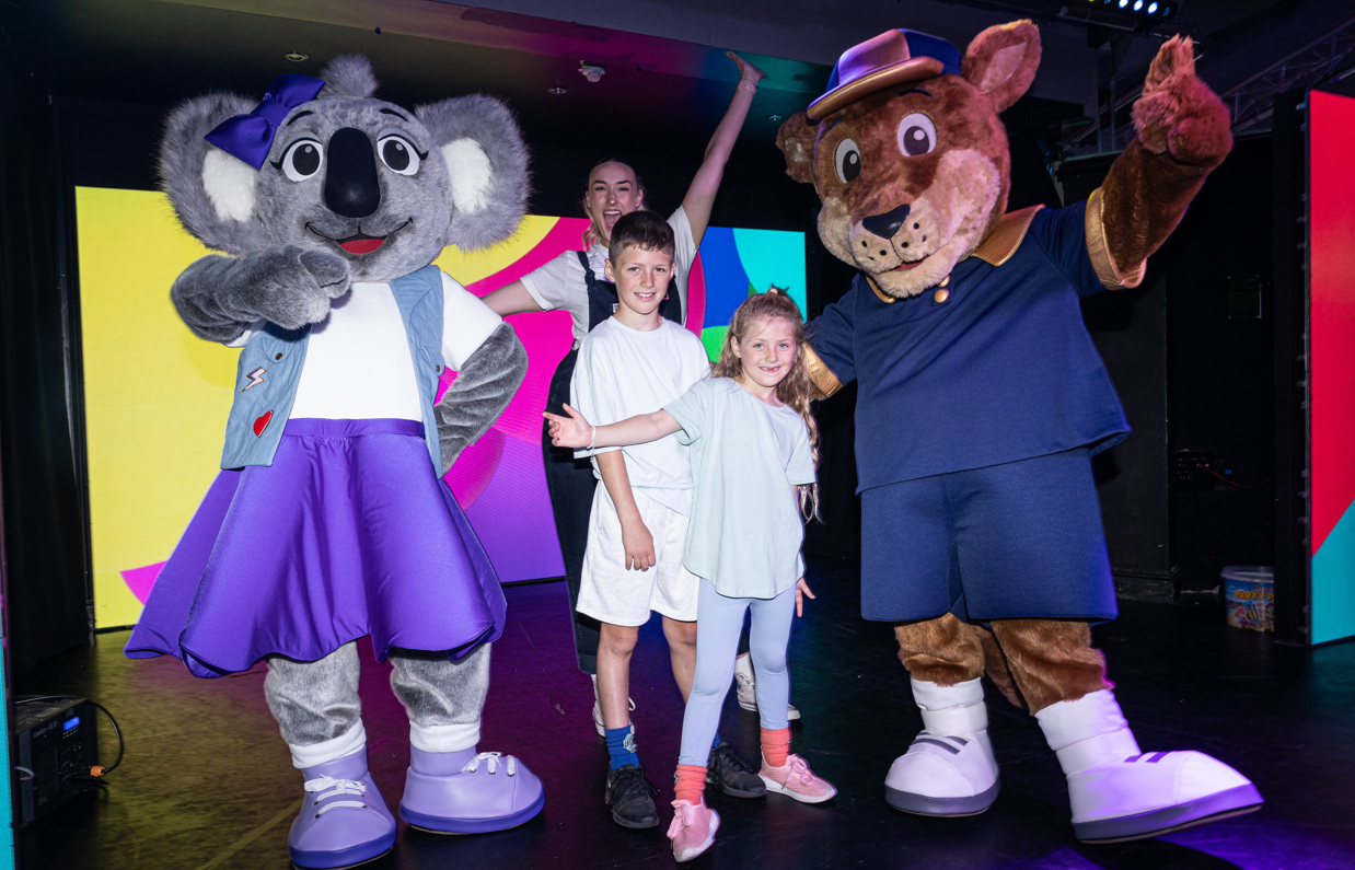 A kangaroo mascot, Boomer, and a koala mascot, Kimi on stage with two young children and an entertainer