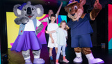 A kangaroo mascot, Boomer, and a koala mascot, Kimi on stage with two young children and an entertainer