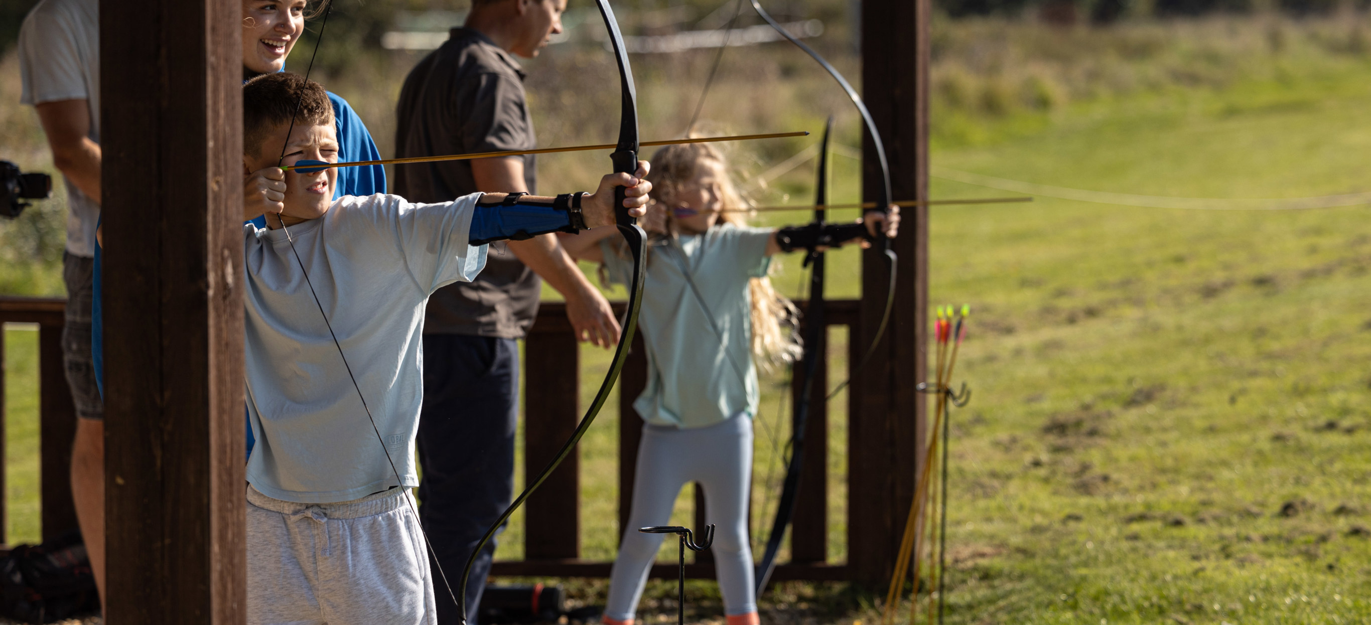 Two young children in a field with activity leaders aiming archery bows at targets