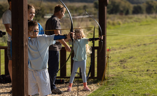 Two young children in a field with activity leaders aiming archery bows at targets