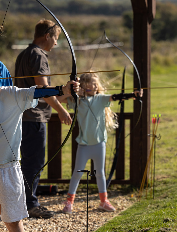 Two young children in a field with activity leaders aiming archery bows at targets