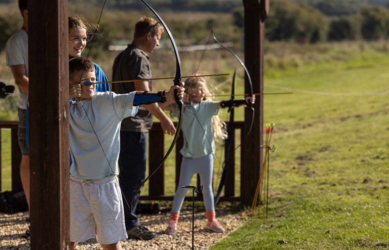 Two young children in a field with activity leaders aiming archery bows at targets