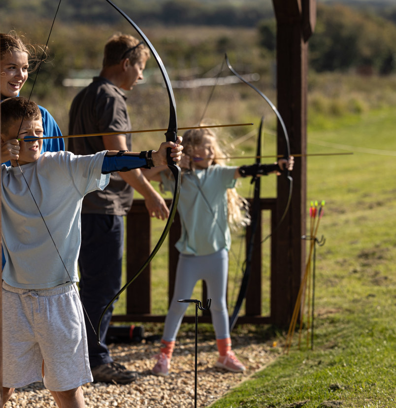 Two young children in a field with activity leaders aiming archery bows at targets