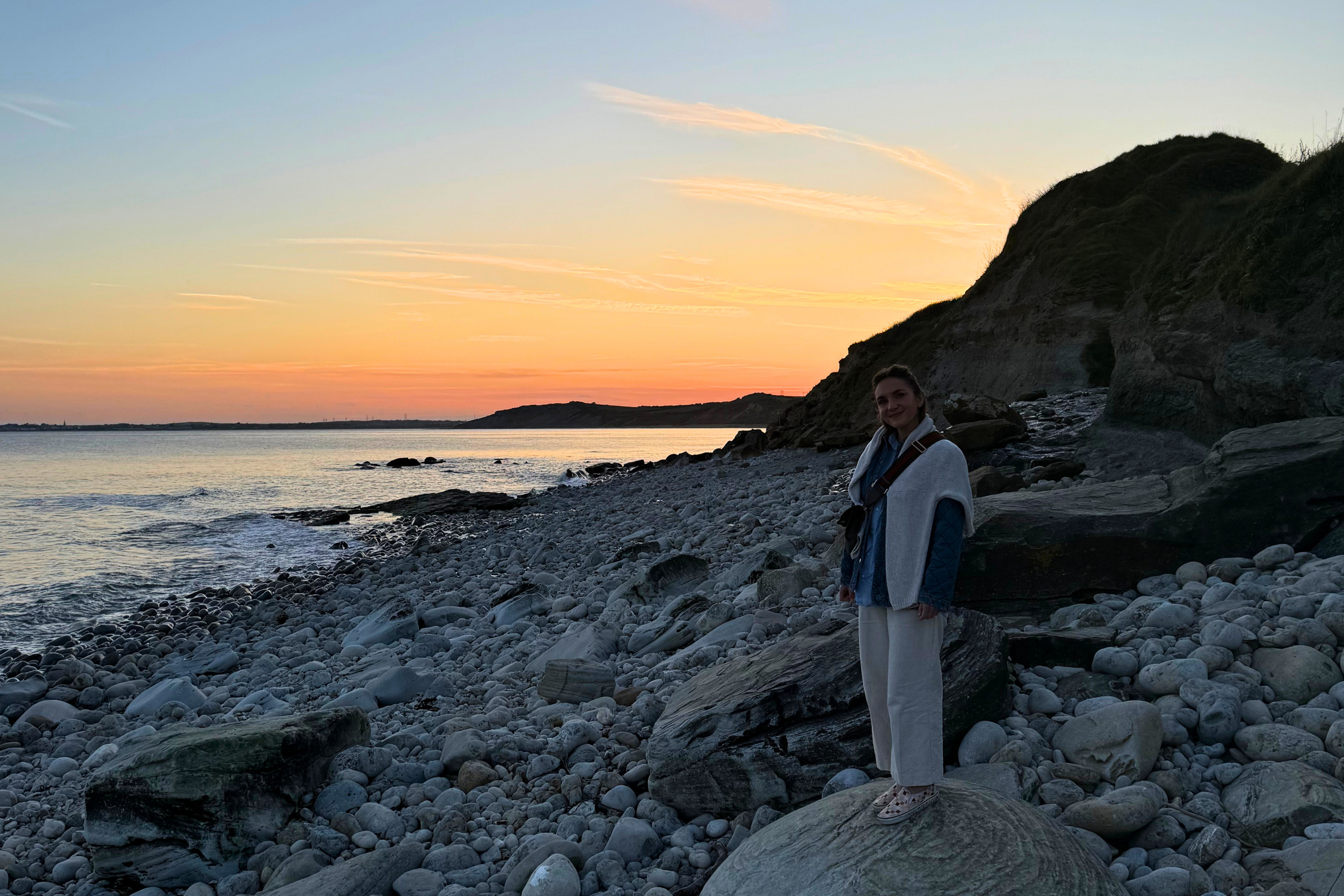 A woman standing on a rock on a pebble beach at sunset with calm waters