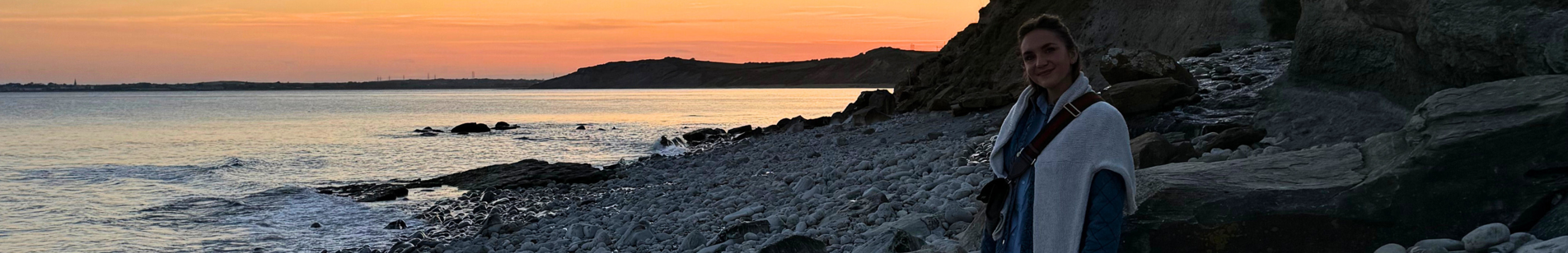 A woman standing on a rock on a pebble beach at sunset with calm waters