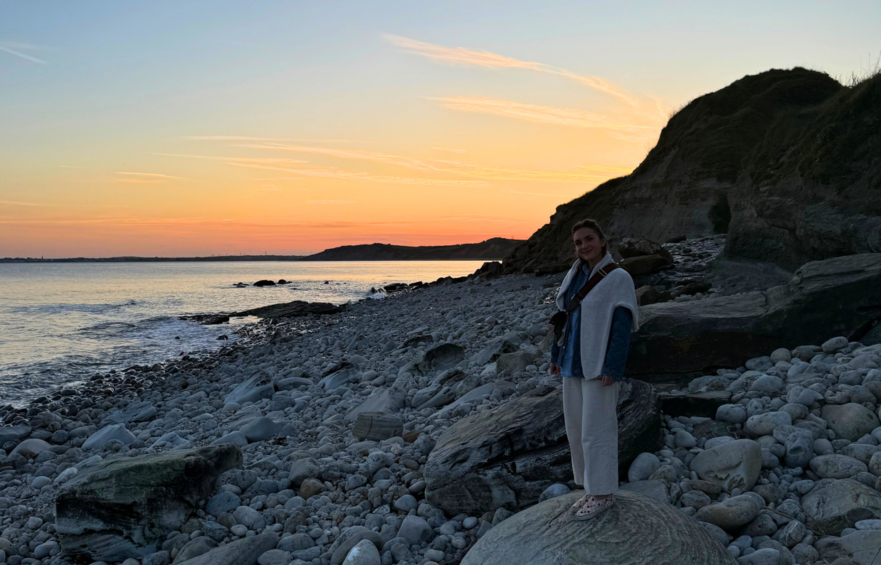 A woman standing on a rock on a pebble beach at sunset with calm waters