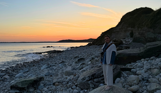 A woman standing on a rock on a pebble beach at sunset with calm waters