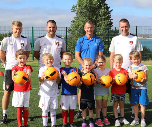 A group of young children with adults all in football / sport gear holding footballs