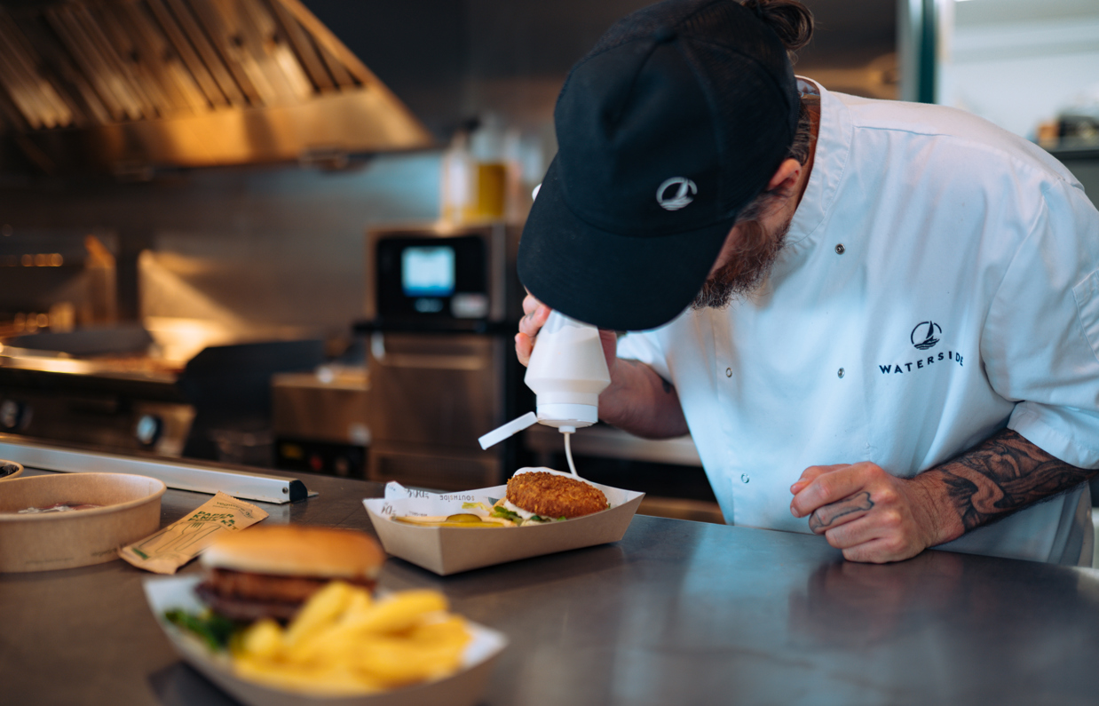 A server at a street food shack applying sauce to a burger with other street food items in shot