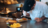 A server at a street food shack applying sauce to a burger with other street food items in shot