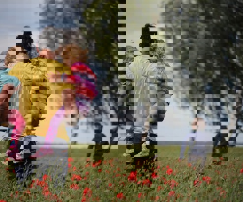 A man carrying two young girls on his hips with a young boy running in front of him in a poppy field with trees in the distance