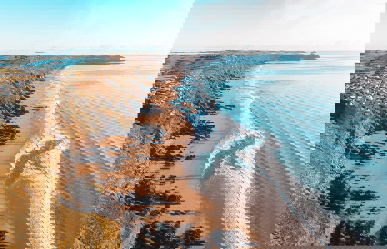 A long stretch of beach with rocky cliffside and blue waves on a sunny, blue sky day