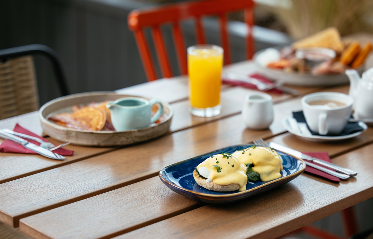 A plate of pancakes with bacon and syrup, a full English and an eggs benedict on a table with various breakfast drinks