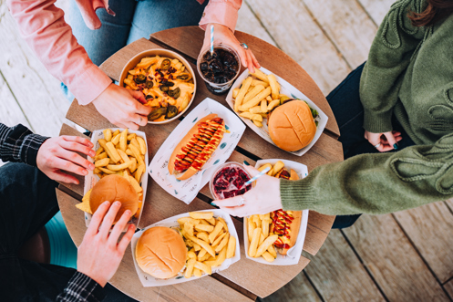 A selection of street food burgers and hot dogs with drinks on a table with three people sat around it