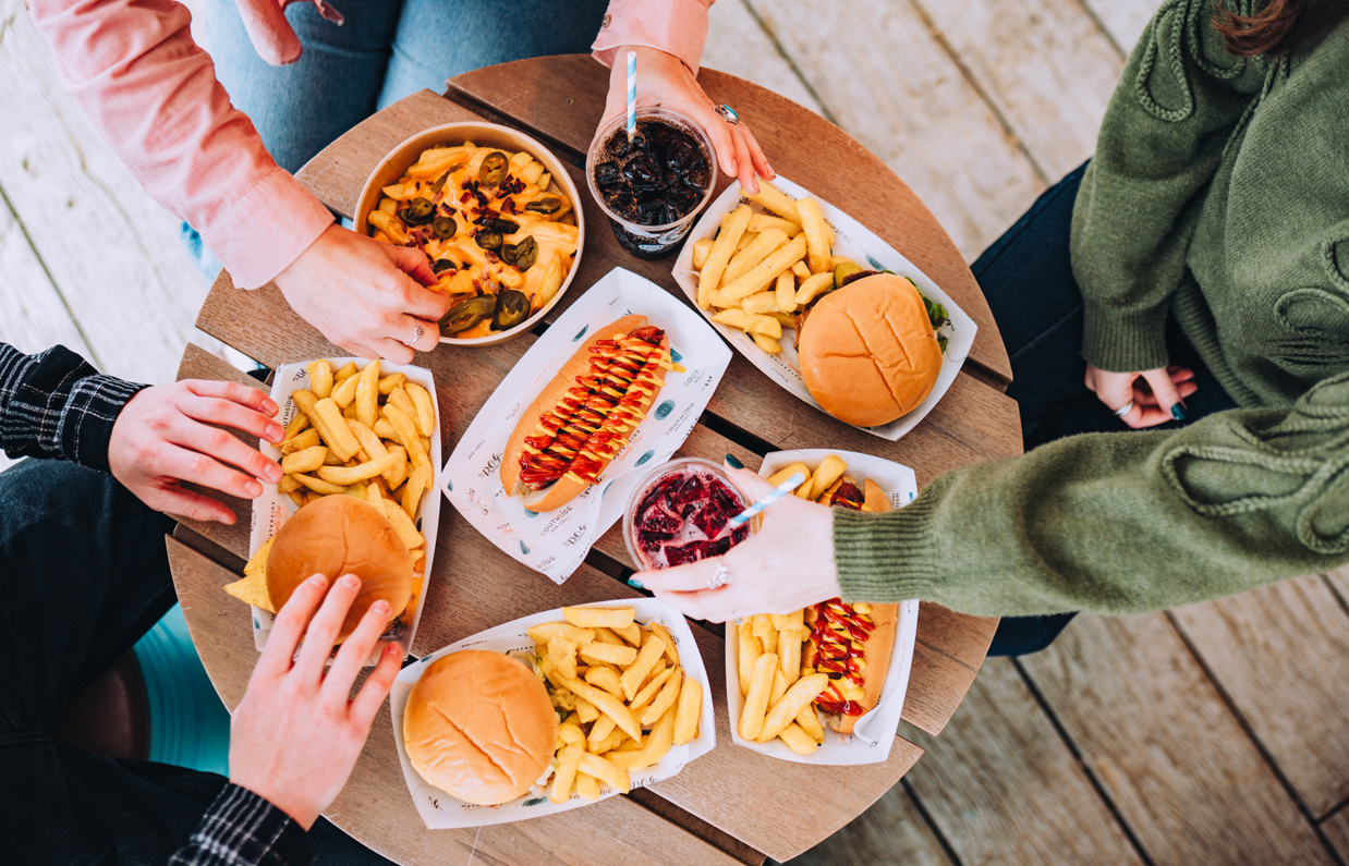 A selection of street food burgers and hot dogs with drinks on a table with three people sat around it