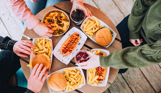 A selection of street food burgers and hot dogs with drinks on a table with three people sat around it