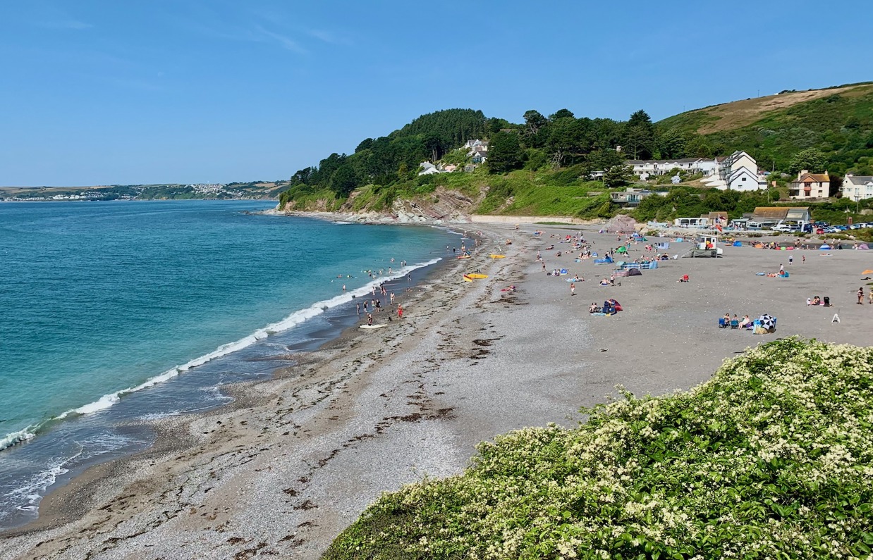 A small shingle beach at the bottom of rolling countryside hills on a sunny, blue sky day