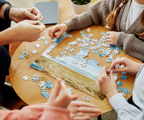 A group of people sat around a table doing a jigsaw puzzle of a beach scene